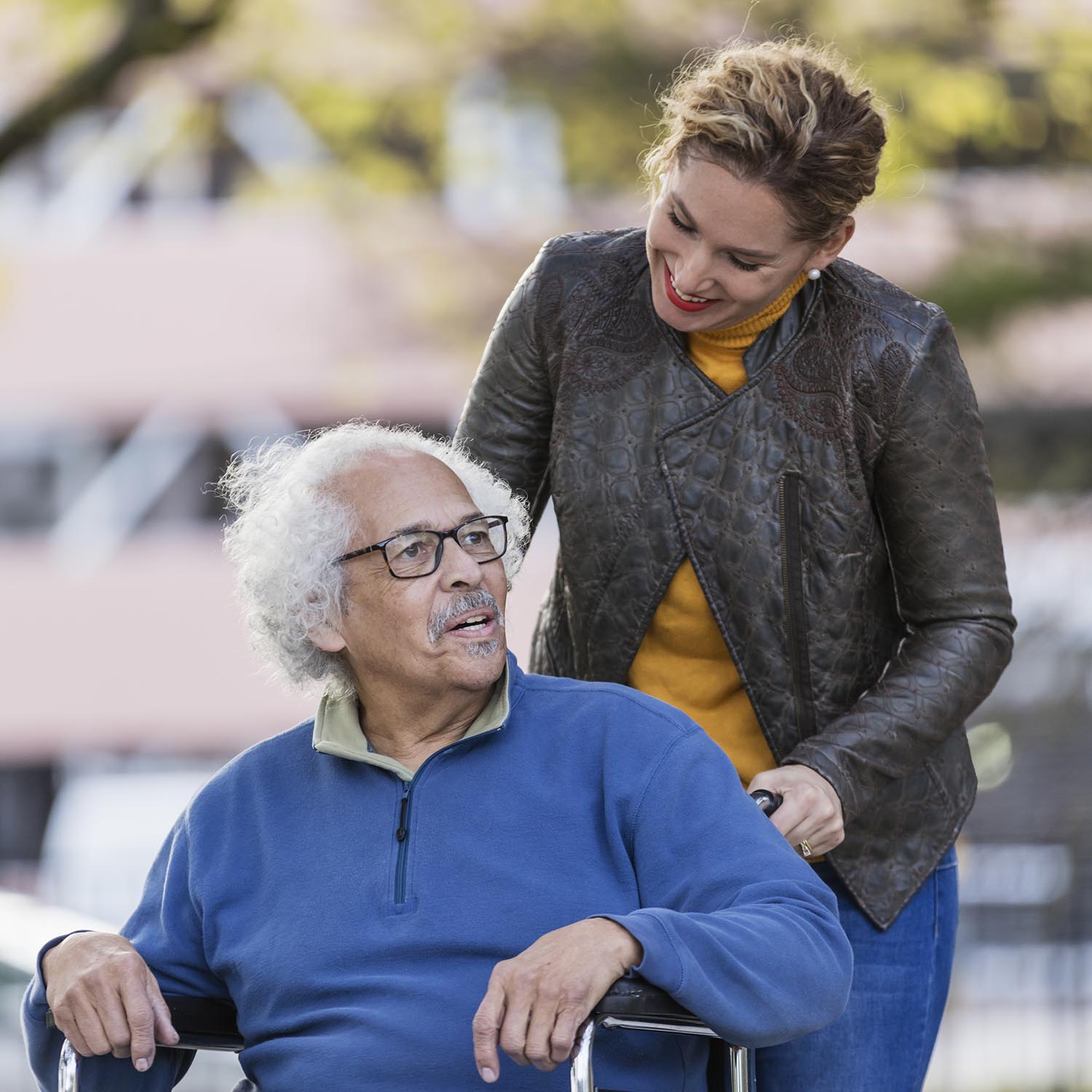 A senior Hispanic man in his 80s sitting in a wheelchair, talking to his adult daughter, who is in her 30s, pushing the wheelchair from behind. They are walking in a city park in fall, building out of focus in the background.