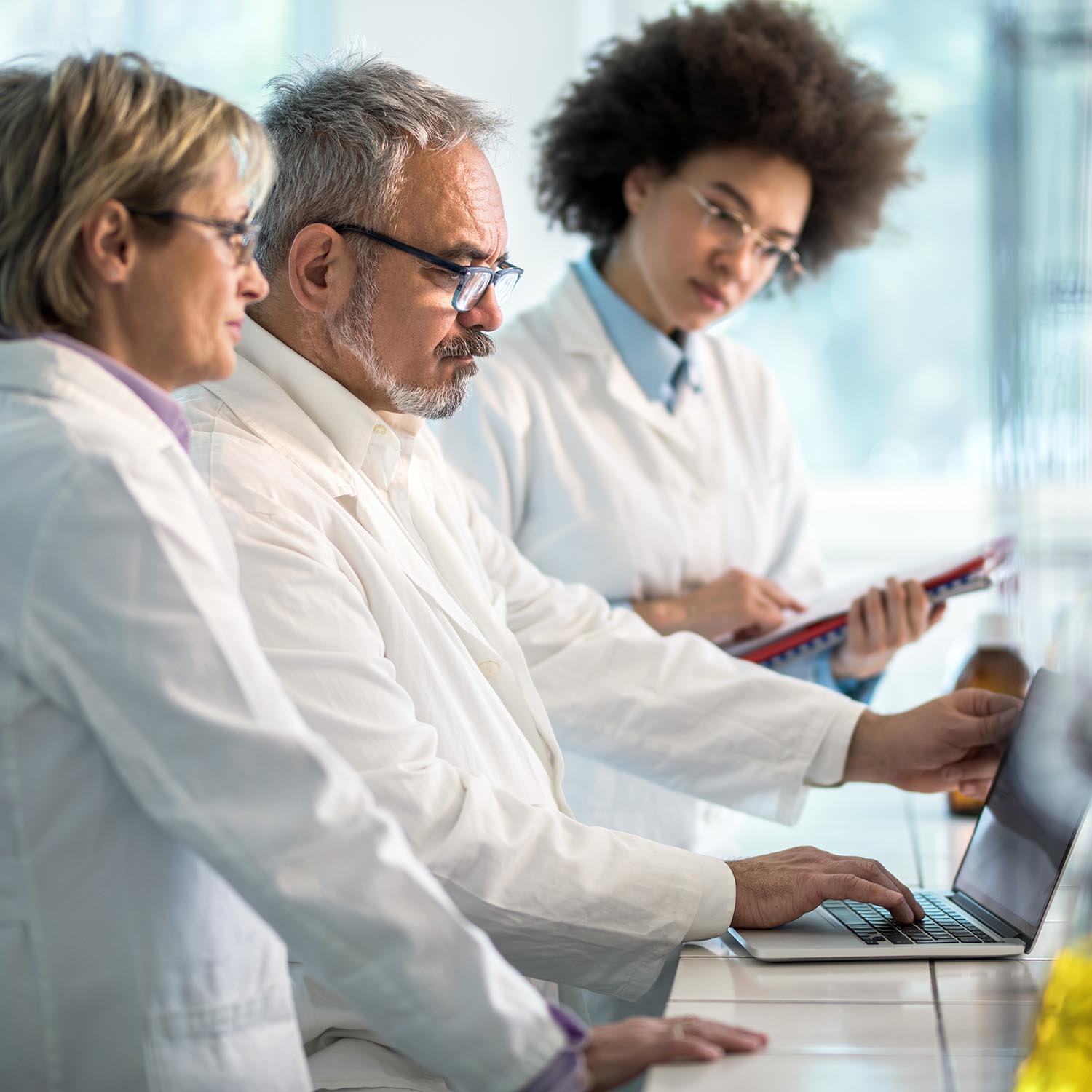 Three biochemists working on laptop in a laboratory. Focus is on mature man typing on laptop.