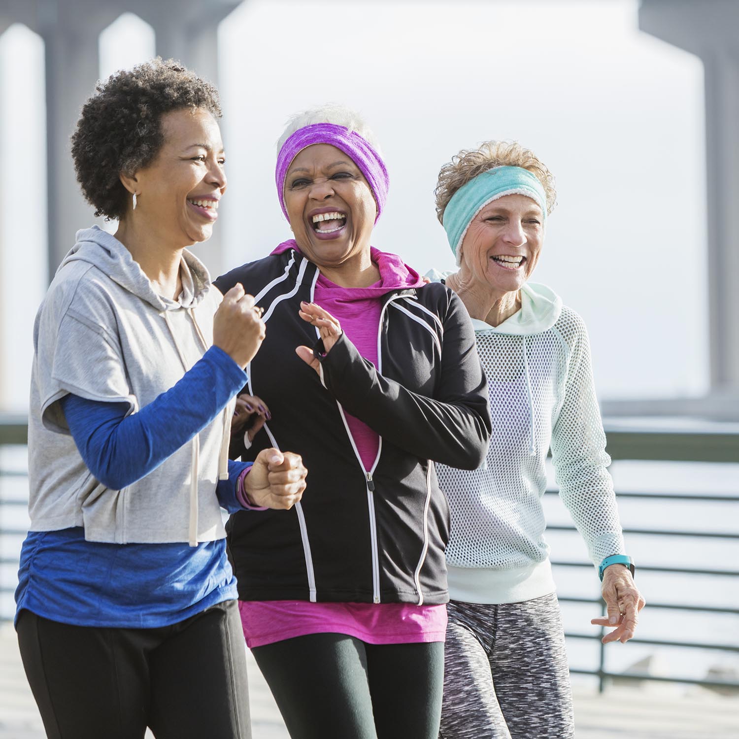 Three multi-ethnic women exercising together, power walking side by side on a city waterfront. The African-American woman with black hair is in her 50s. Her friends are senior women in their 60s.