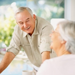 An elderly man and woman sit at a table next to a window talking, with mugs on the table before them.