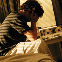 A man sits on the edge of his bed, tiredly holding his head in his hands.